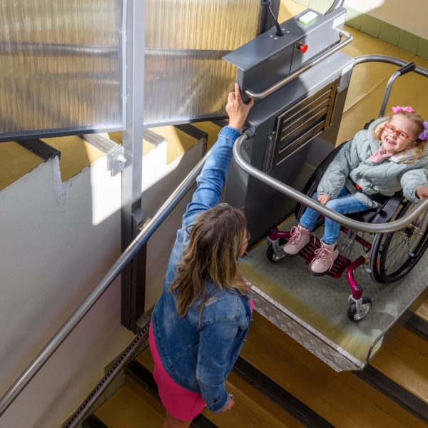 Mother with a young child living with cerebral palsy using electric wheelchair lift to access public building. Special lifting platform for wheelchair users. Disability stairs lift facility.